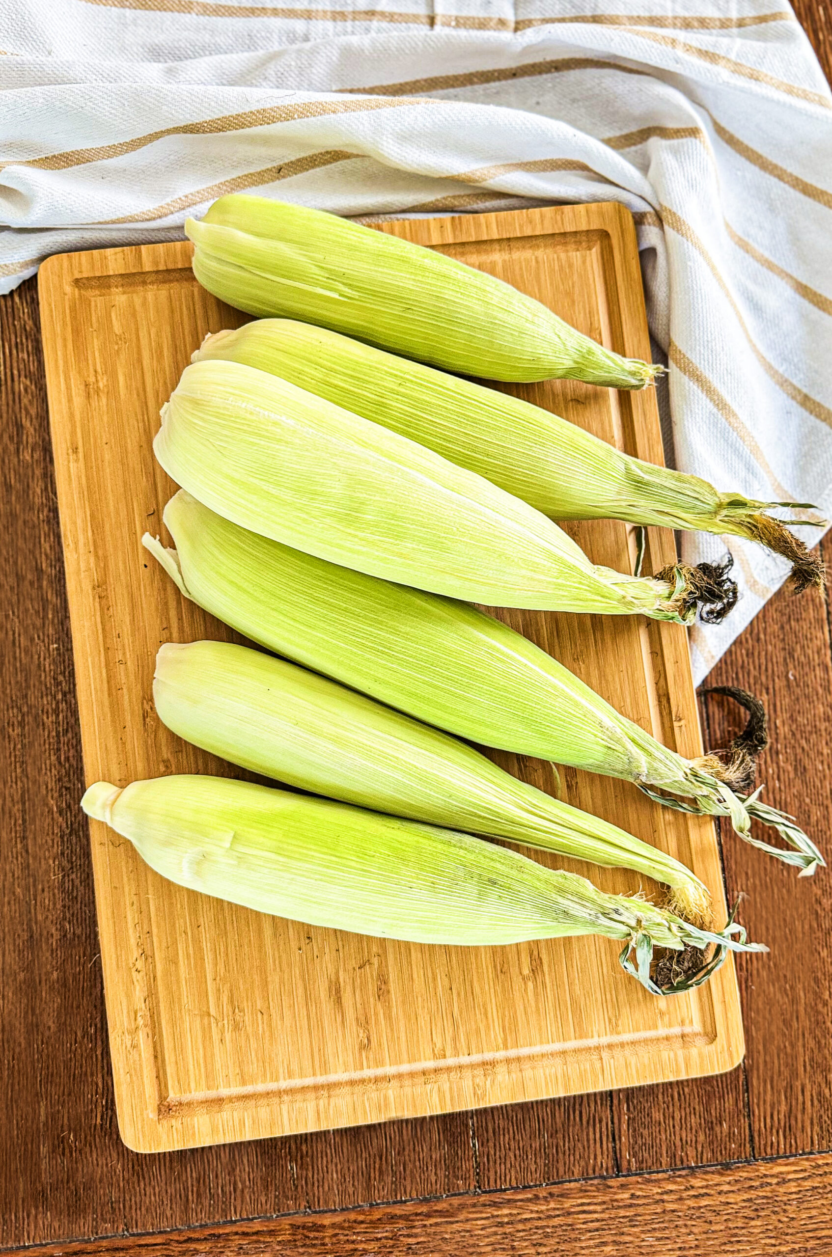 A row of corn cobs in their husks on a wooden cutting board.