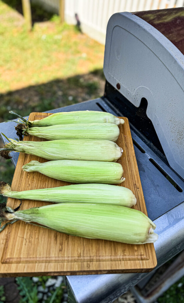 Corn in their husk on a wooding cutting board getting ready to go on the grill.