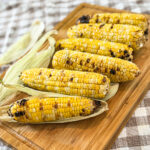 Grilled corn on a wooden cutting board on a checked table cloth.