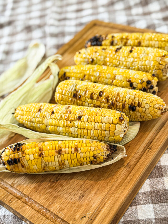 Grilled corn on a wooden cutting board on a checked table cloth.