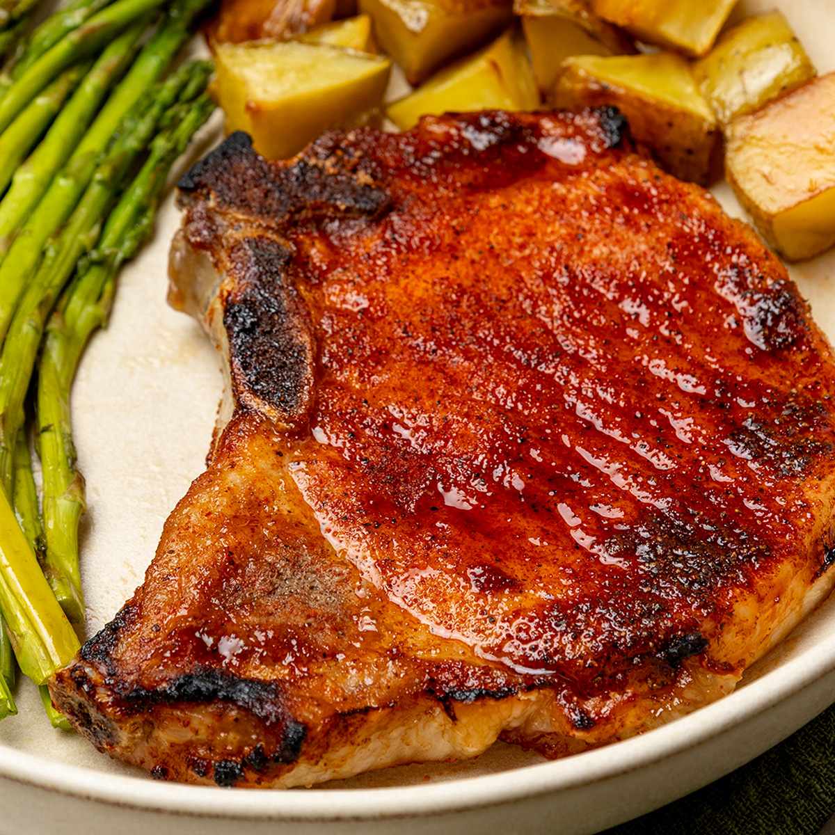 Browned oven baked bone-in pork chop on a white plate with roasted potatoes and asparagus.