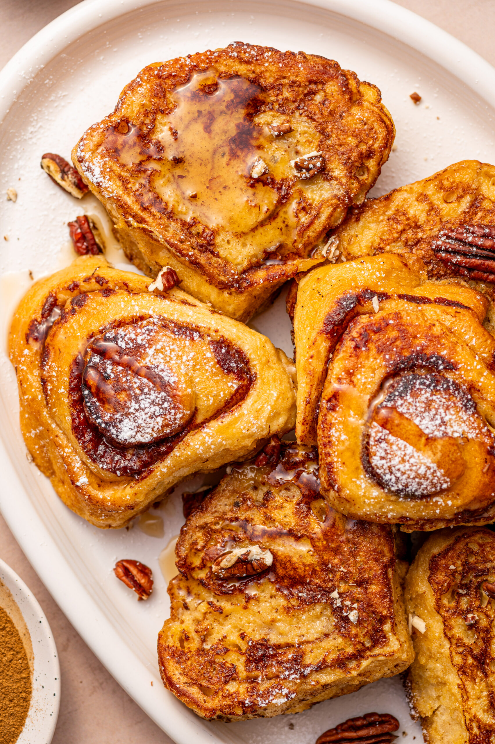A white plate of cinnamon roll French toast with maple syrup and pecans.