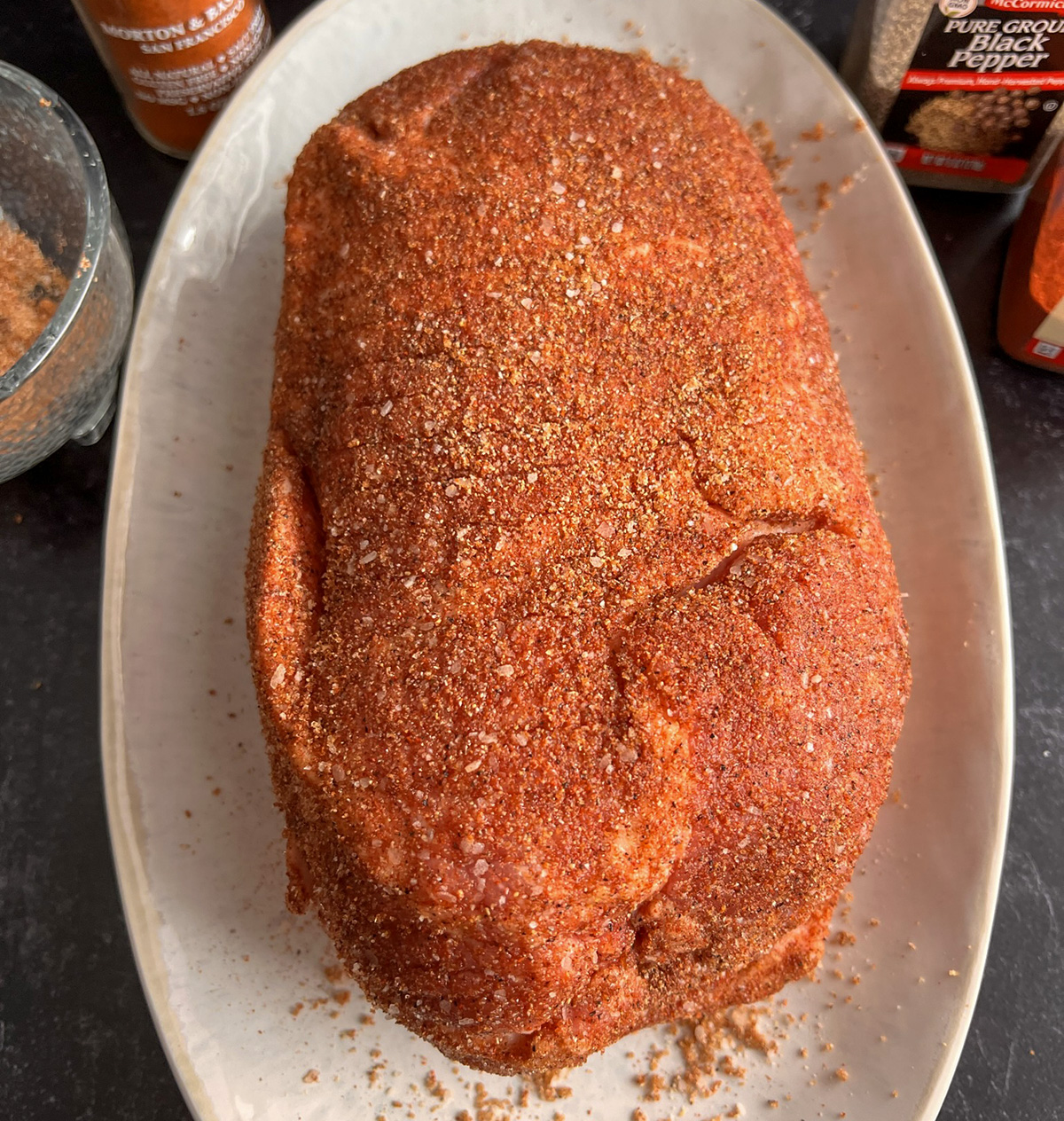 Uncooked boneless pork butt coated with a reddish-brown dry rub on a white oval platter.