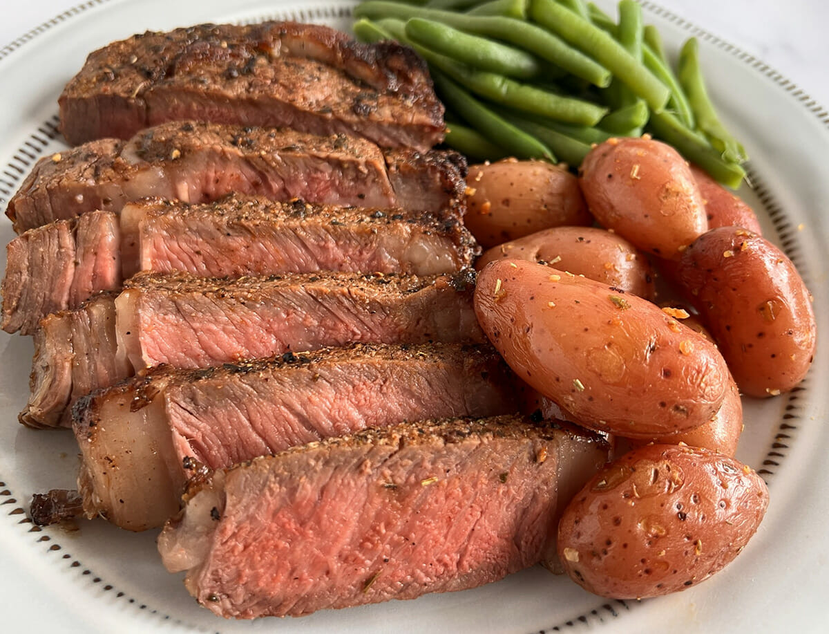 A plate of sliced ribeye steak made in an air fryer, served with potatoes and green beans.