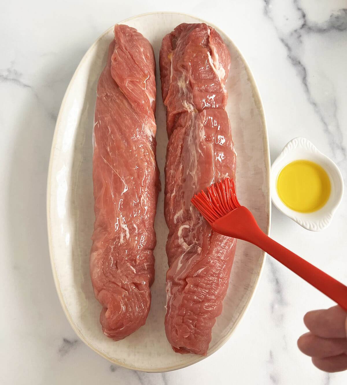 A plate of 2 pork tenderloins being coated with olive oil.