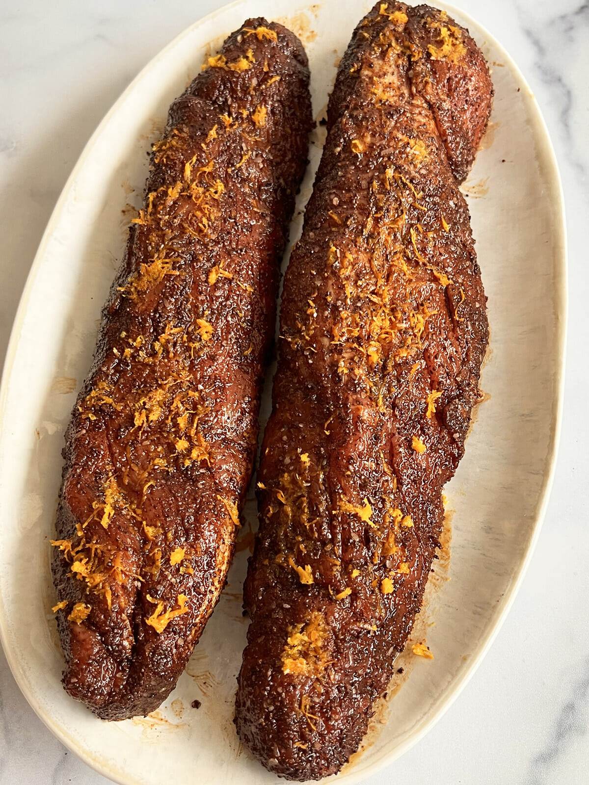 Two pork tenderloins on a plate seasoned with a coffee rub and orange zest on top.