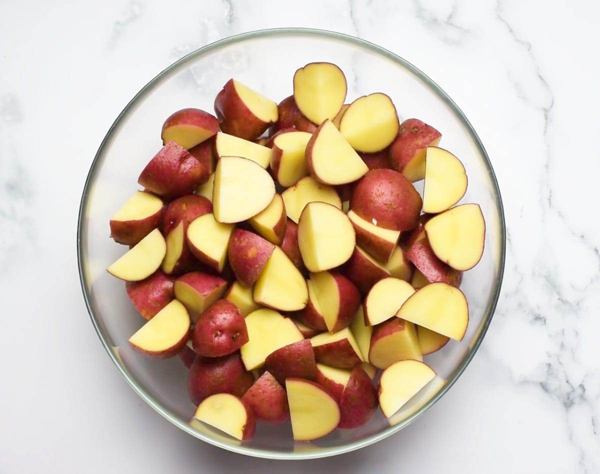 A bowl of red potatoes, quartered over a white plate on a white marble surface.
