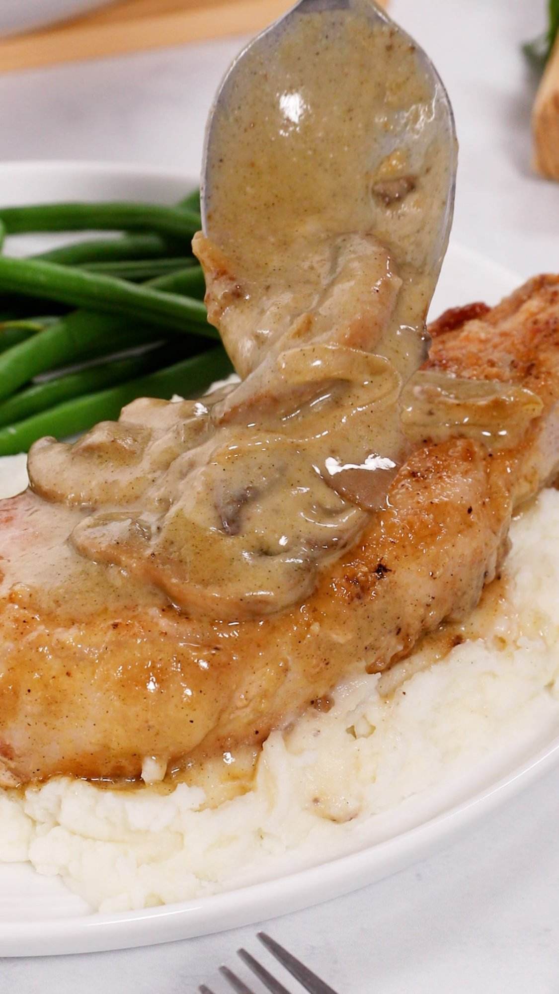 A pork chop atop mashed potatoes being topped with an onion and mushroom gravy for smothered pork chops.