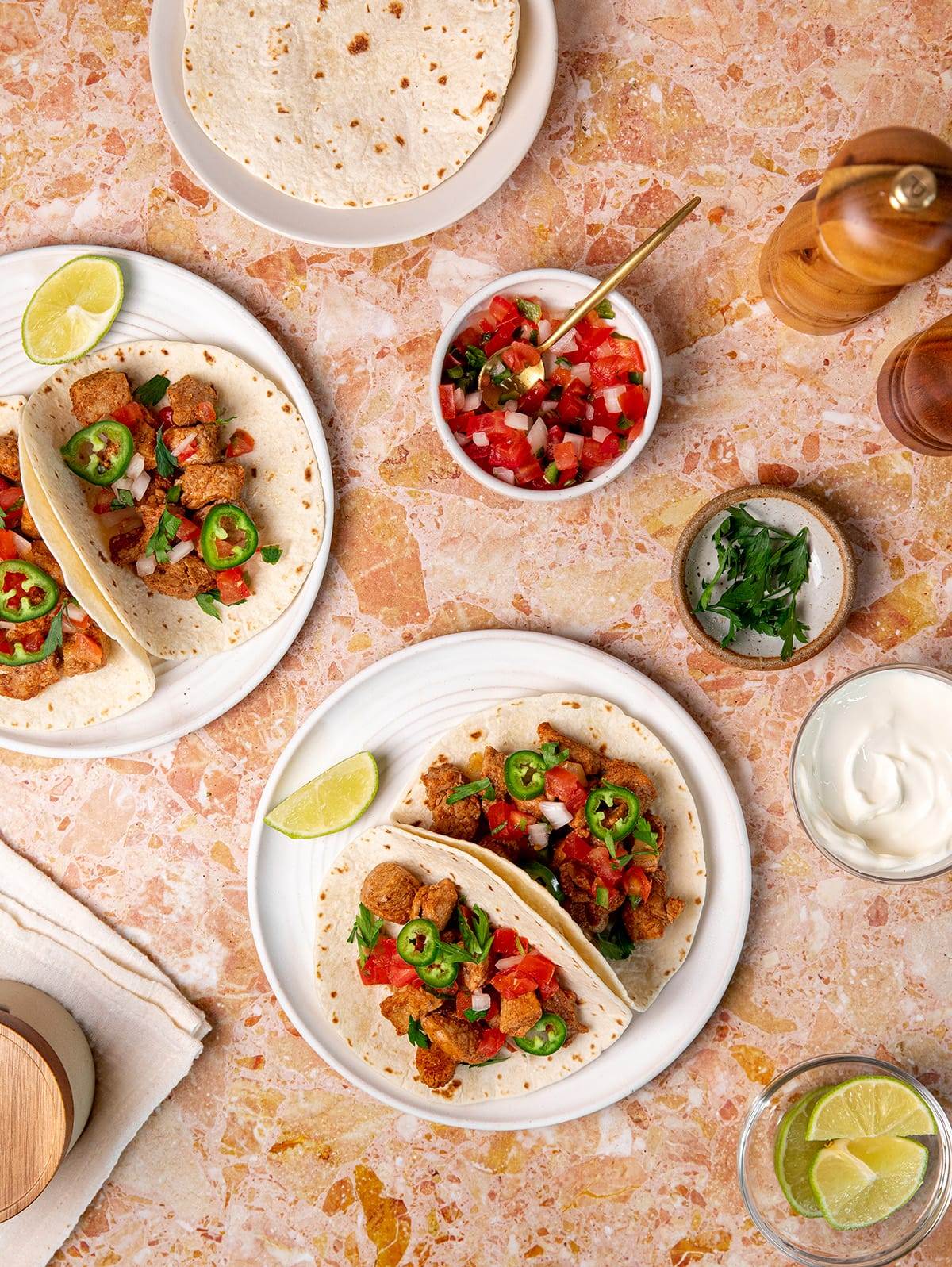 A table setting with pork tacos and toppings including pico de gallo, cilantro and sour cream.