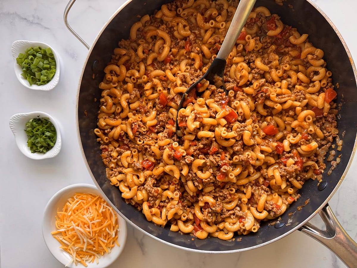 A skillet with ground beef taco pasts and toppings and cheese on the side.