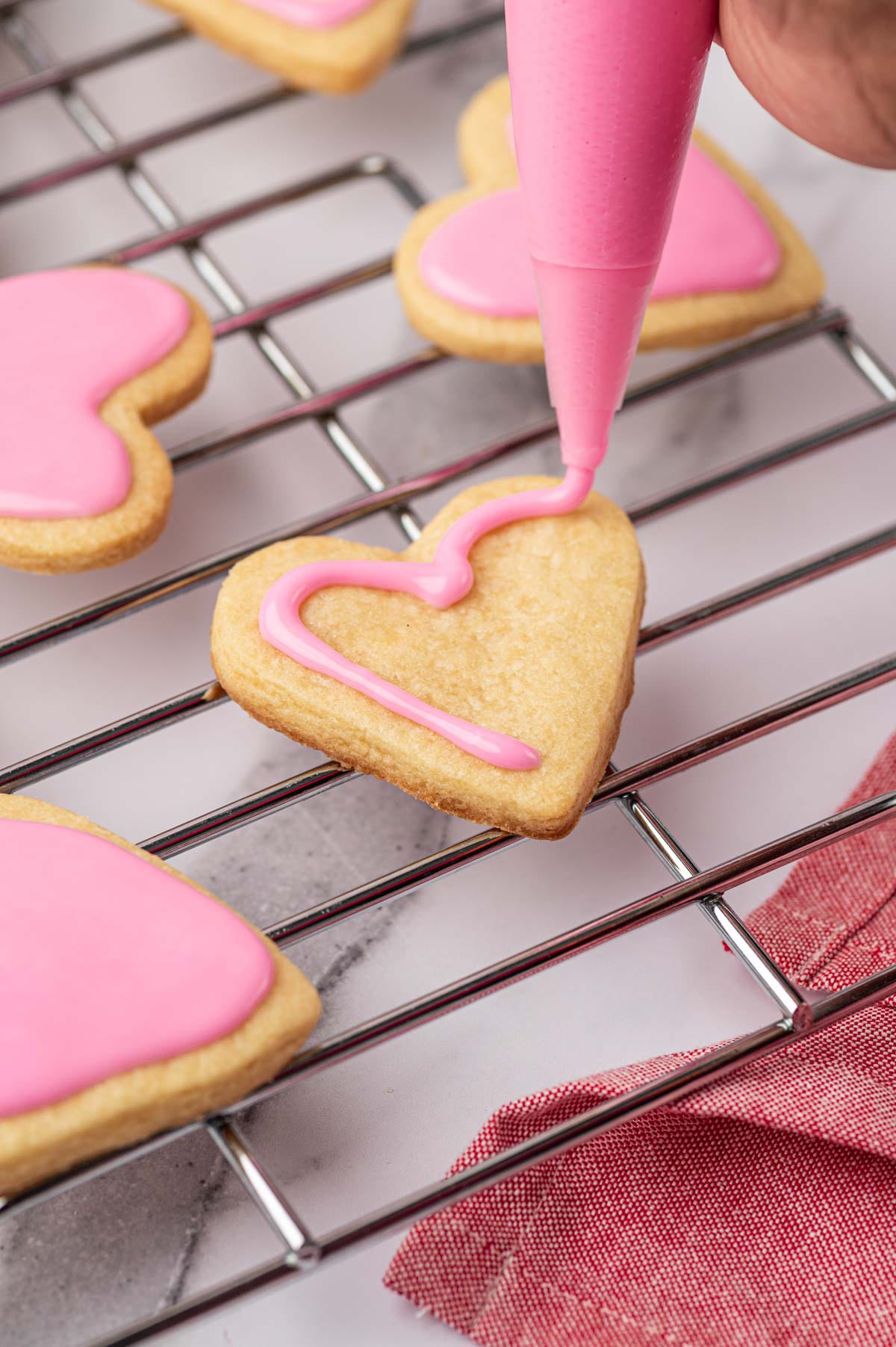 Heart-shaped sugar cookies on a cooling rack getting frosted with a piping bag.