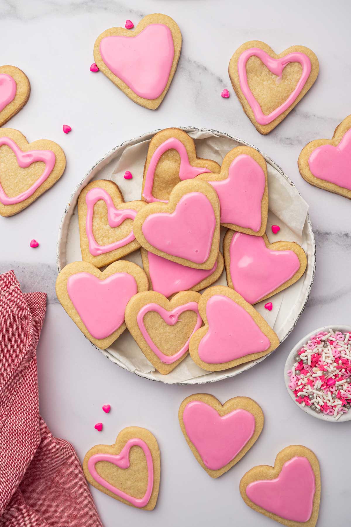 Hero shot of heart-shaped sugar cookies on a white plate with a bowl of sprinkles.