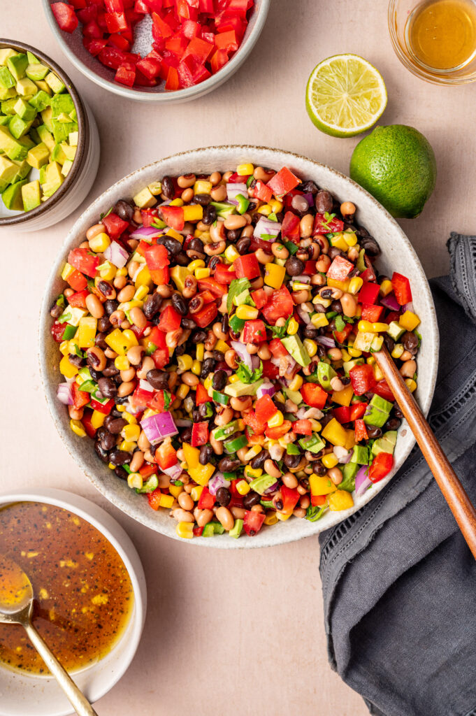 A large white bowl of cowboy caviar with a wooden spoon and dressing on the side.