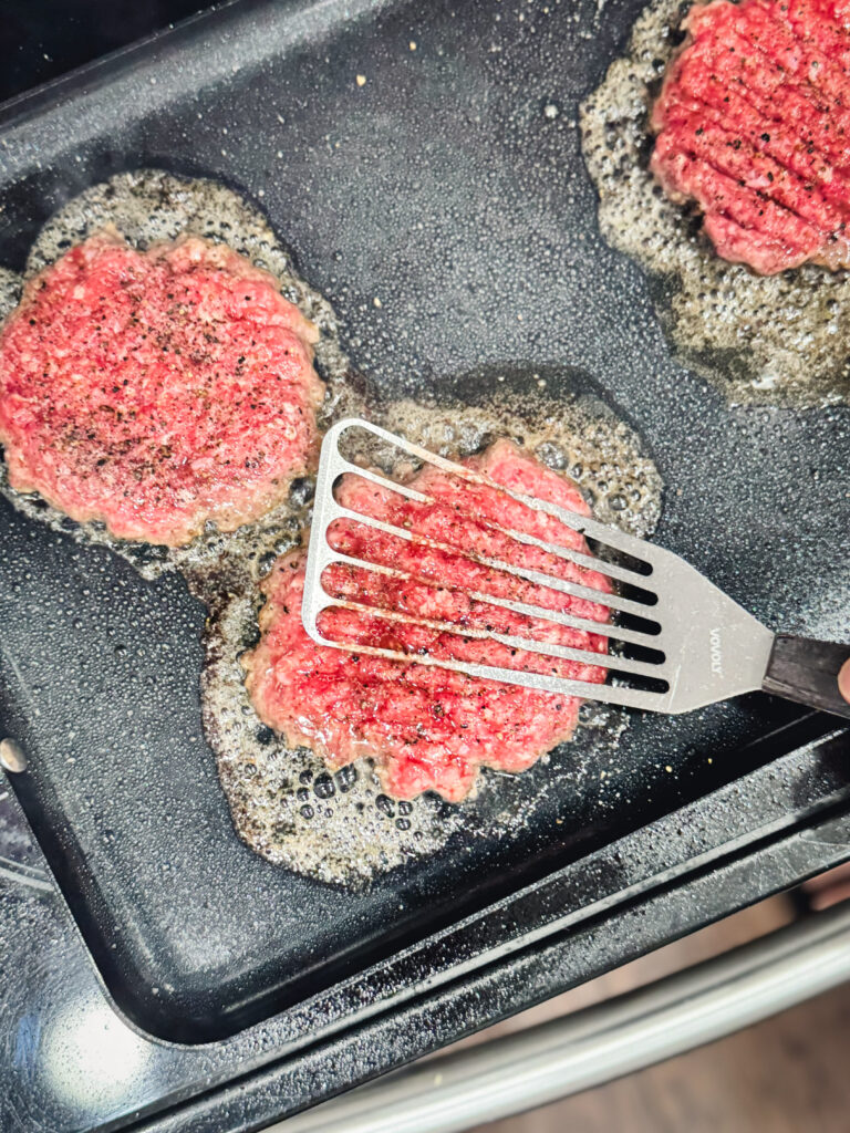 Pressing ground beef patties on a griddle with a spatula.