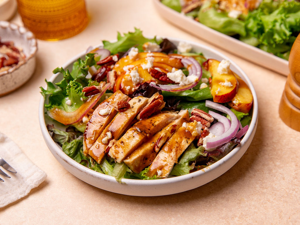 Grilled peach and chicken salad in a white bowl with onion, goat cheese, and dressing.