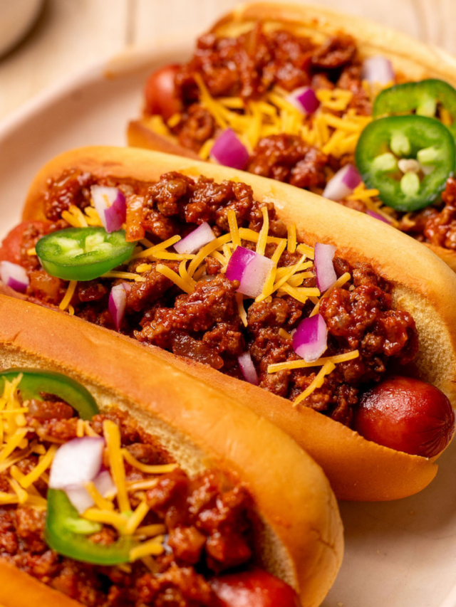 Three loaded chili dogs on a serving dish with shredded cheese and sliced jalapenos.