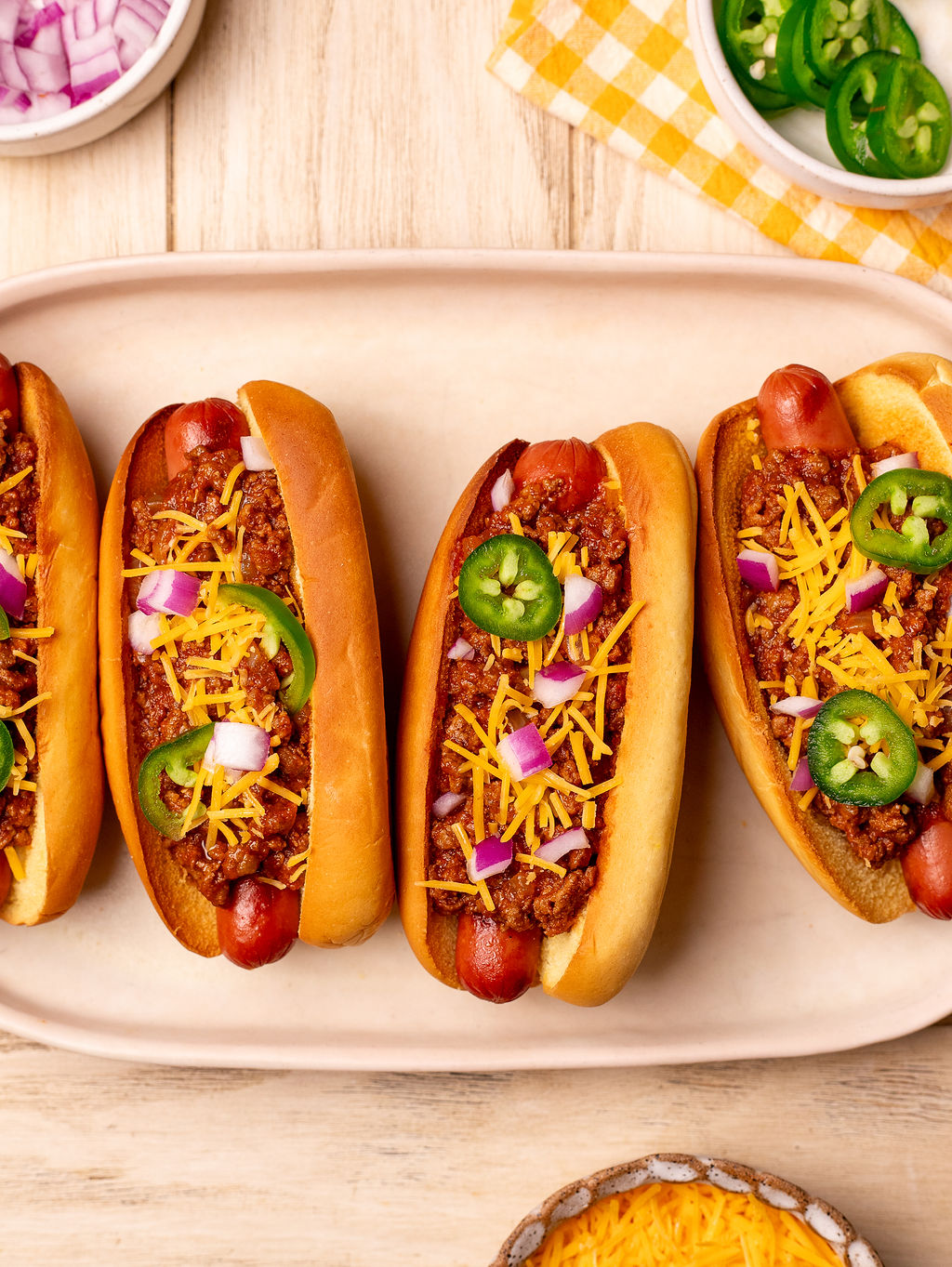 Four chili dogs on a serving plate with shredded cheese, onion, and jalapeno as toppings.