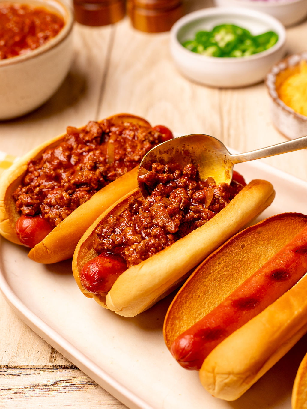 Adding chili to three hot dogs in a row on a serving plate.