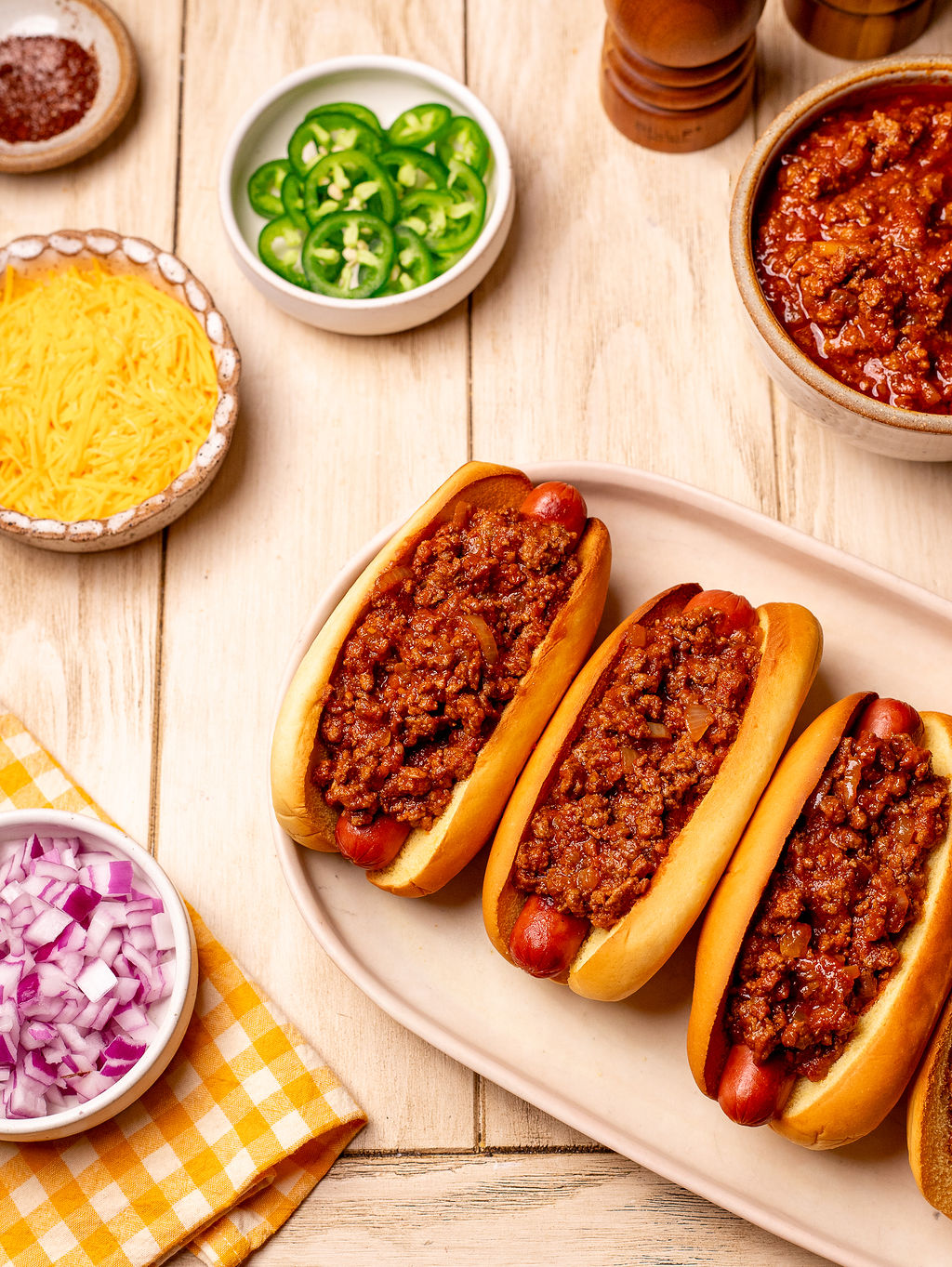 Three chili dogs in a row on a serving plate with ingredients surrounding it.