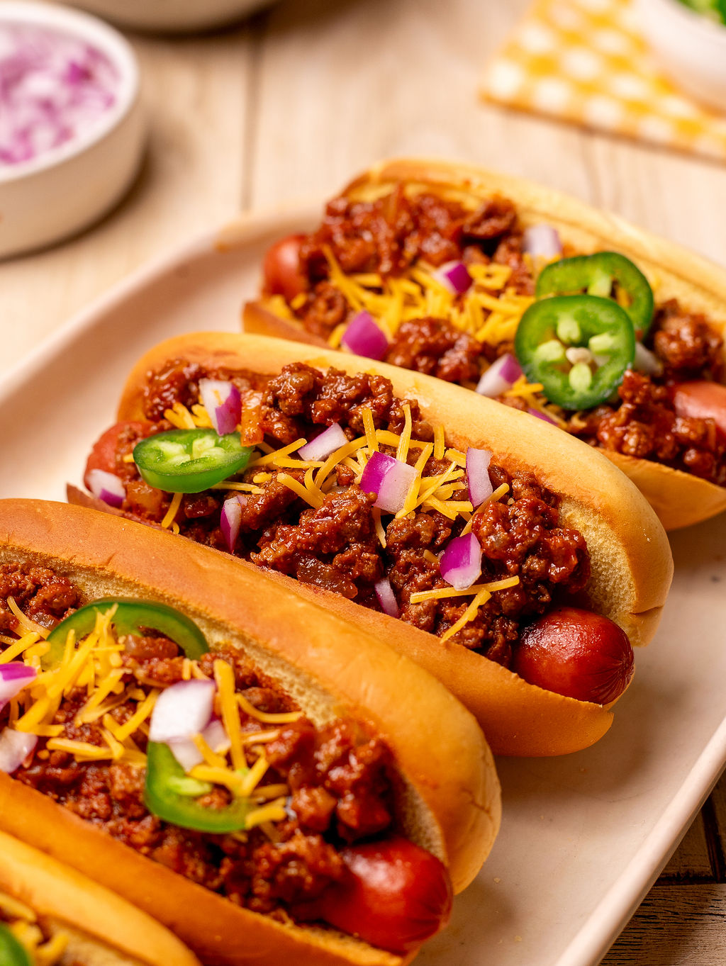Three loaded chili dogs on a serving dish with shredded cheese and sliced jalapenos.