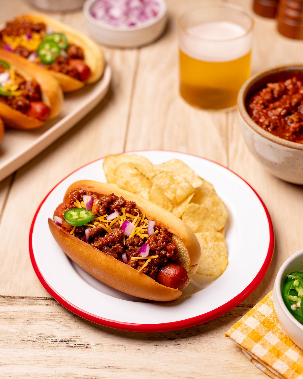 A chili dog with shredded cheese and jalapeno with a side of potato chips on a red and white plate.