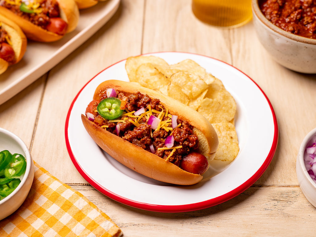 A chili dog with shredded cheese and jalapeno with a side of potato chips on a plate.