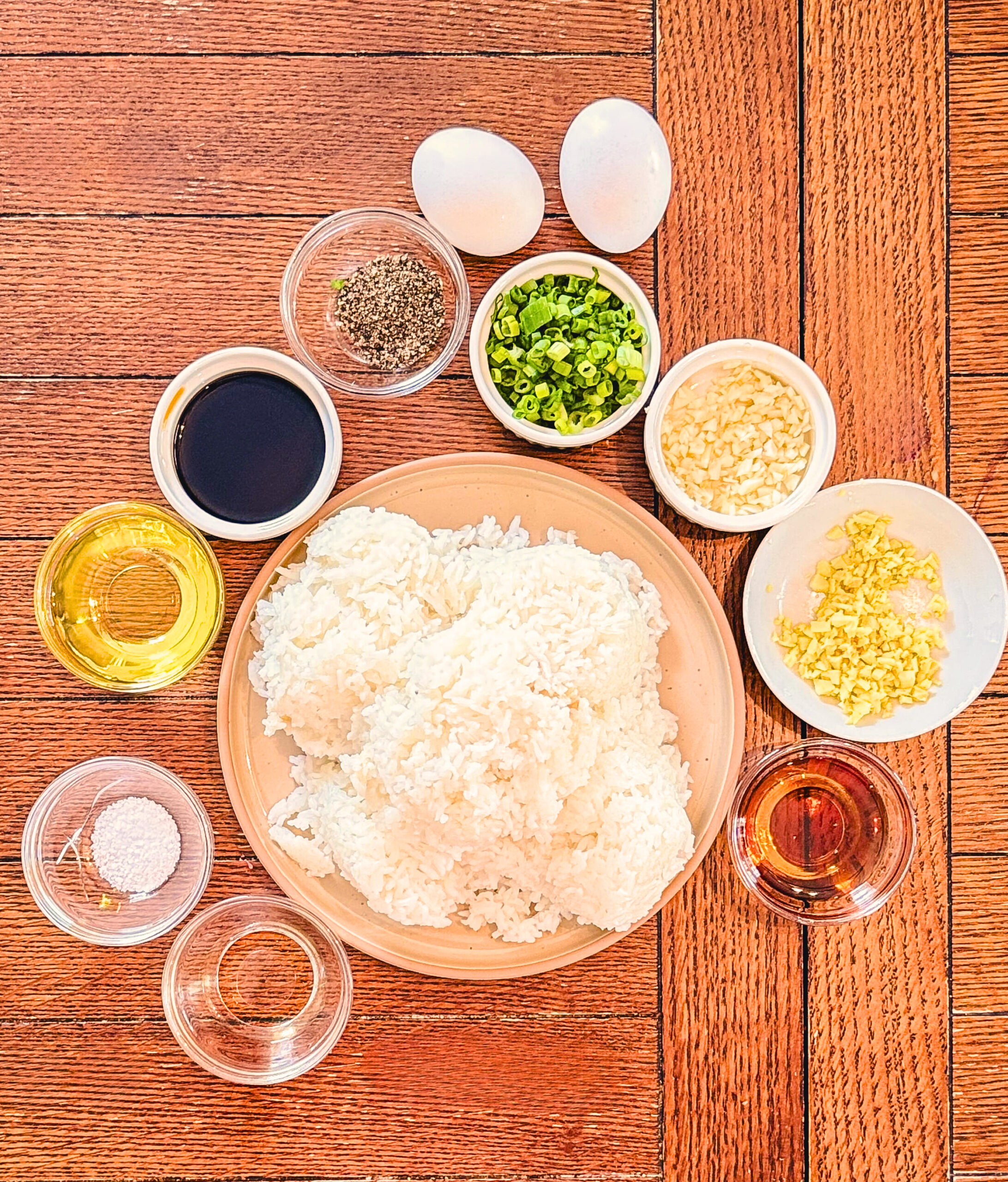 Overhead view of ingredients for fried rice, including a plate of cooked white rice, two eggs, chopped green onions, minced garlic and ginger, black pepper, soy sauce, sesame oil, rice vinegar, salt, and cooking oil, all arranged neatly on a wooden surface.