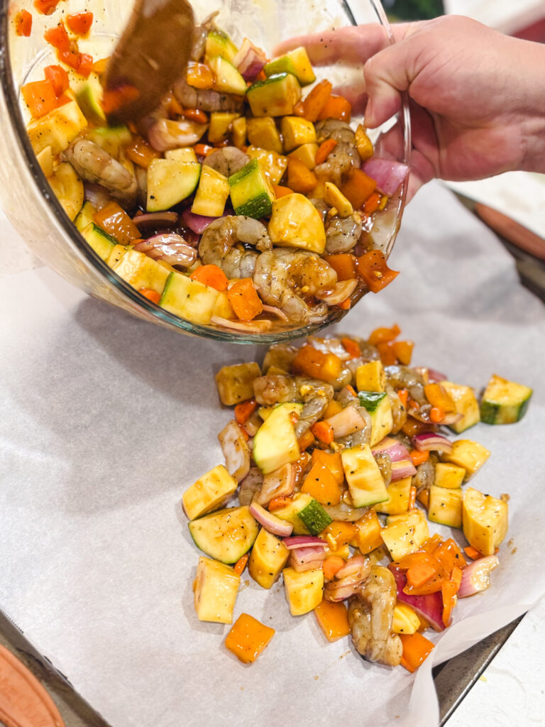 Pouring teriyaki shrimp and veggies on a parchment paper lined baking sheet.