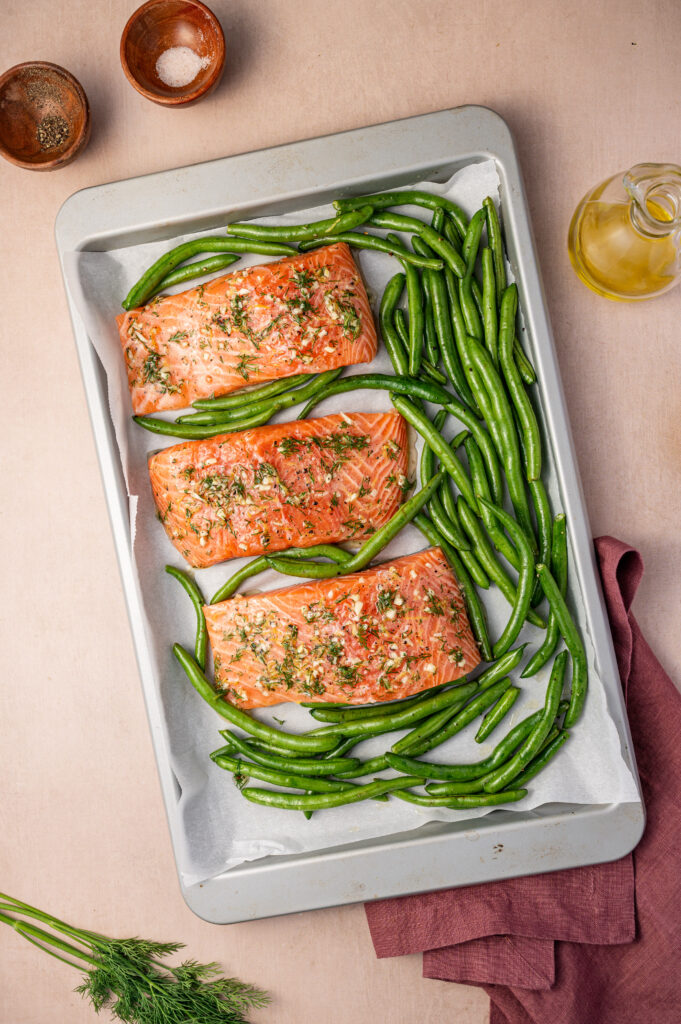 Three lemon dill salmon fillets on a sheet pan with green beans and lemon slices, ready to go in the oven.