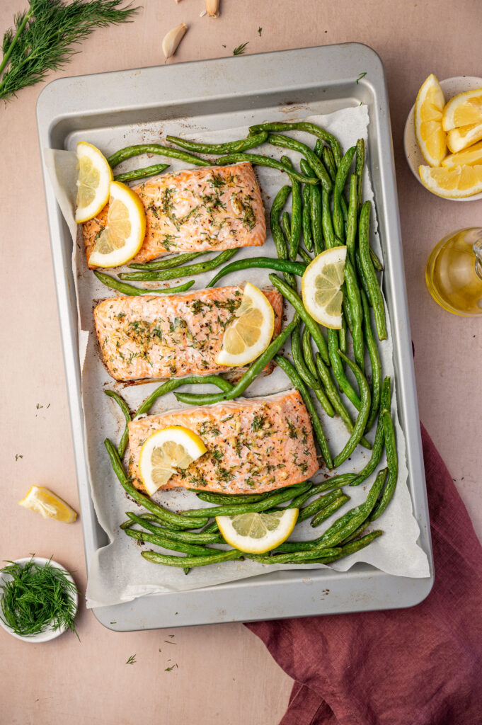 Three lemon dill salmon fillets on a sheet pan with green beans and lemon slices.