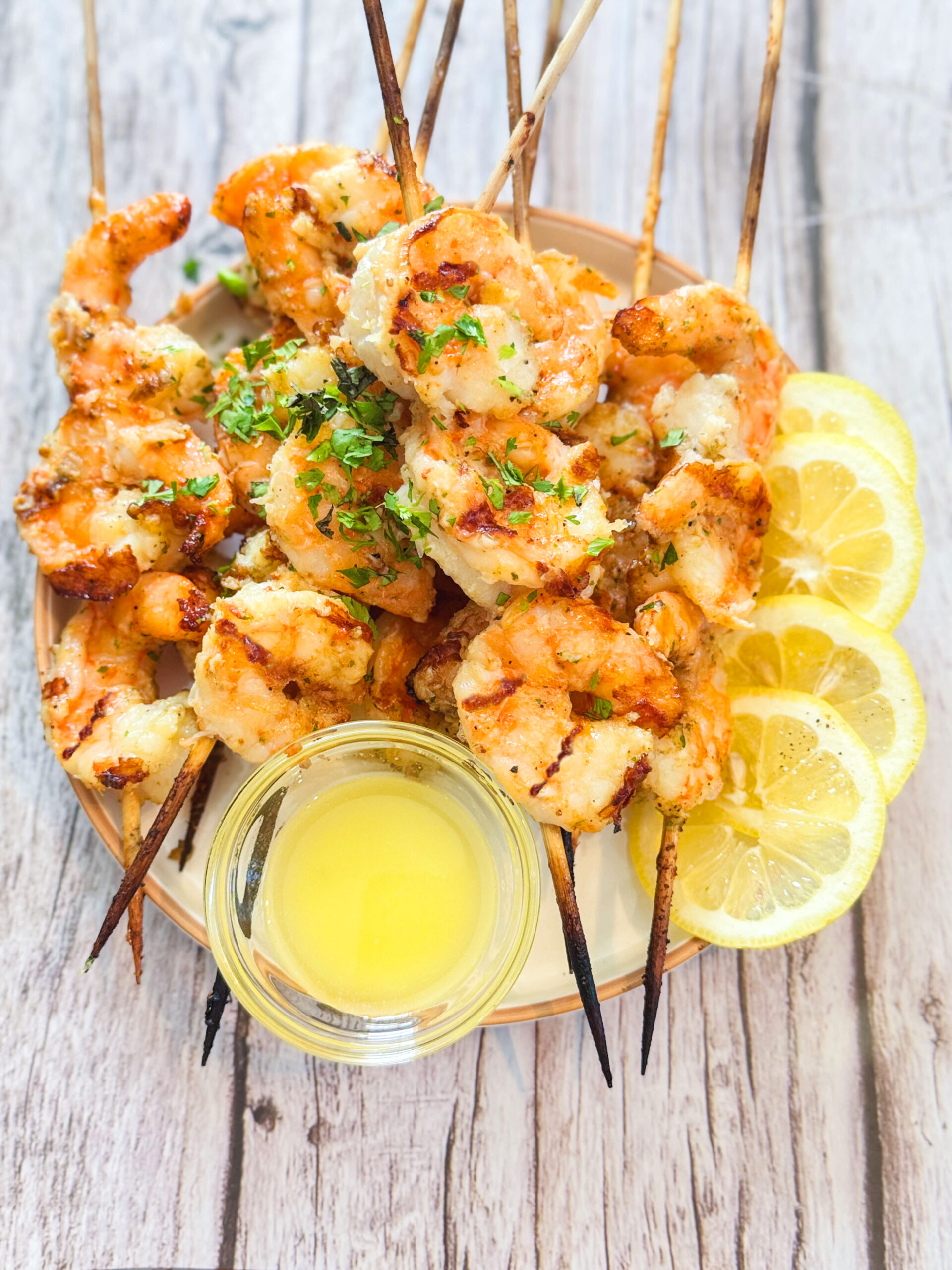 Grilled shrimp skewers on a plate with garlic butter and sliced lemon on the side.