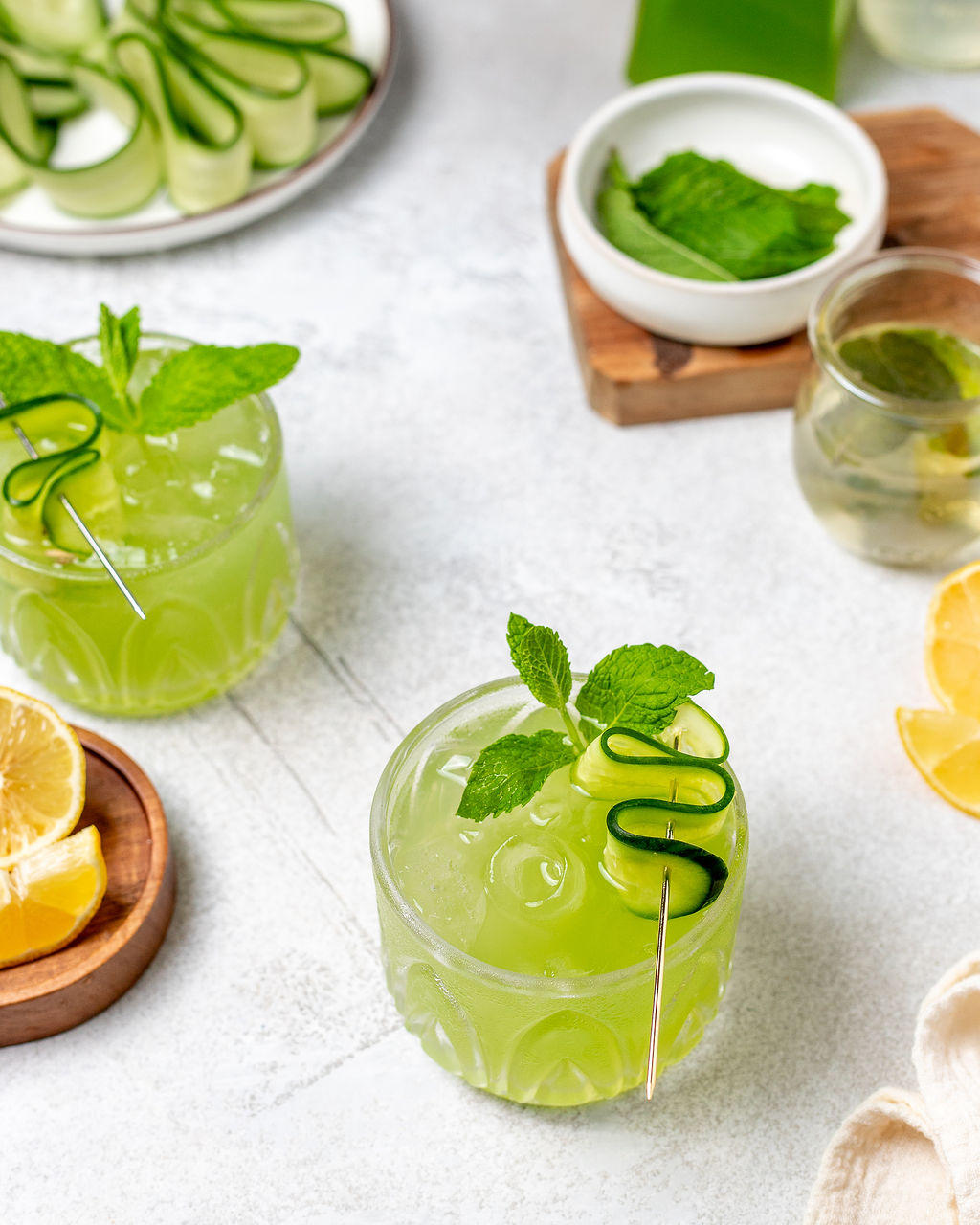 Cucumber coolers in glasses surrounded by ingredients on a white marble surface.