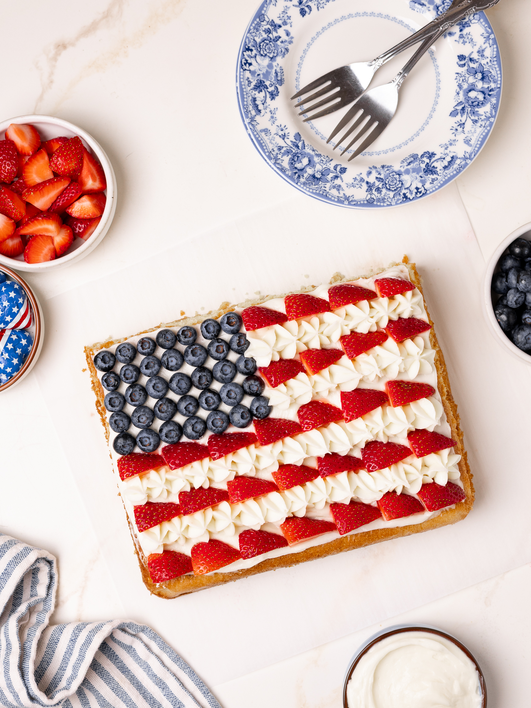 A rectangular American flag cake decorated with blueberries for the stars and sliced strawberries with piped white frosting for the stripes. The cake is surrounded by bowls of fresh berries, whipped cream, and patriotic chocolate candies.