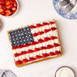 A rectangular American flag cake decorated with blueberries for the stars and sliced strawberries with piped white frosting for the stripes. The cake is surrounded by bowls of fresh berries, whipped cream, and patriotic chocolate candies.