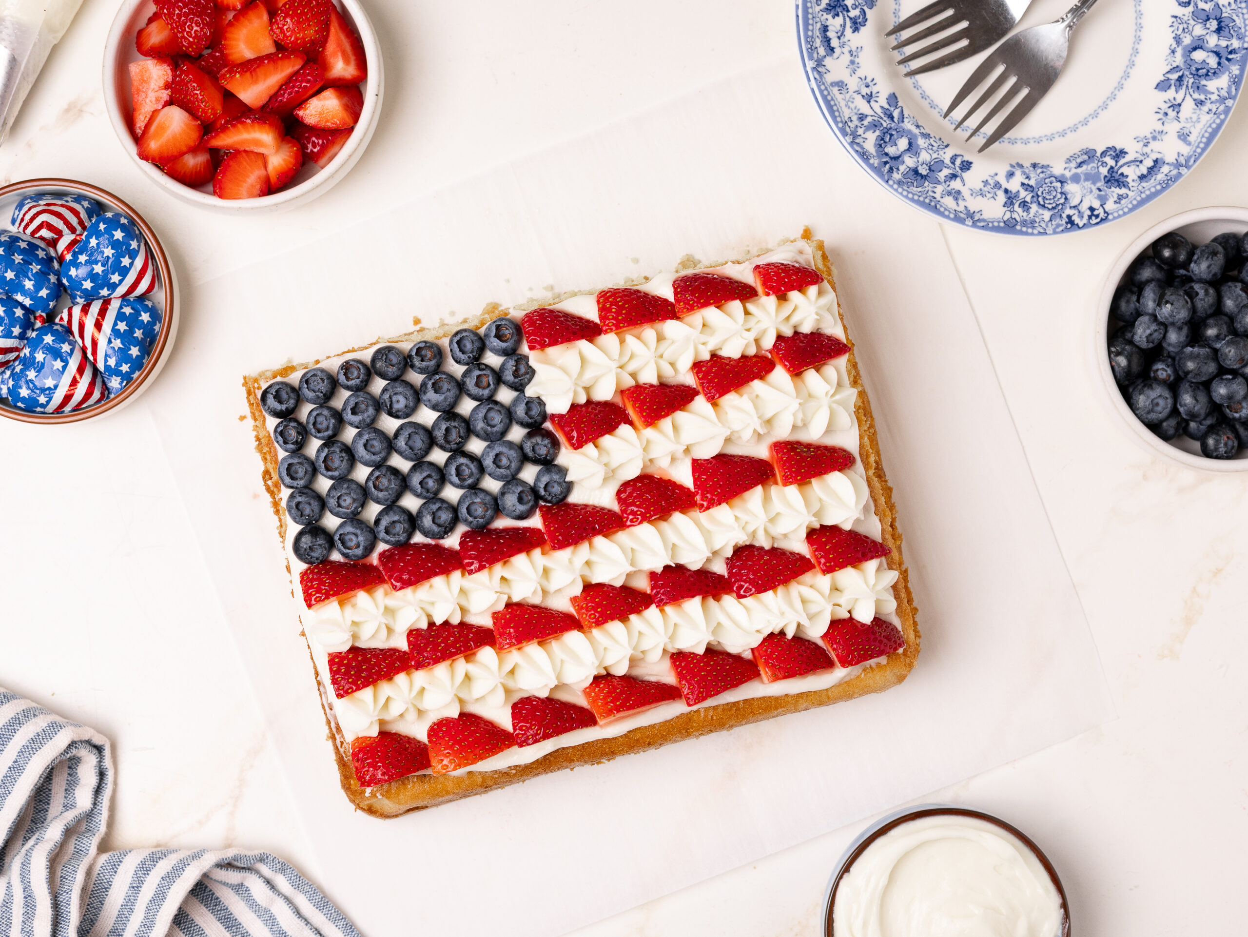 A rectangular American flag cake decorated with blueberries for the stars and sliced strawberries with piped white frosting for the stripes. The cake is surrounded by bowls of fresh berries, whipped cream, and patriotic chocolate candies.