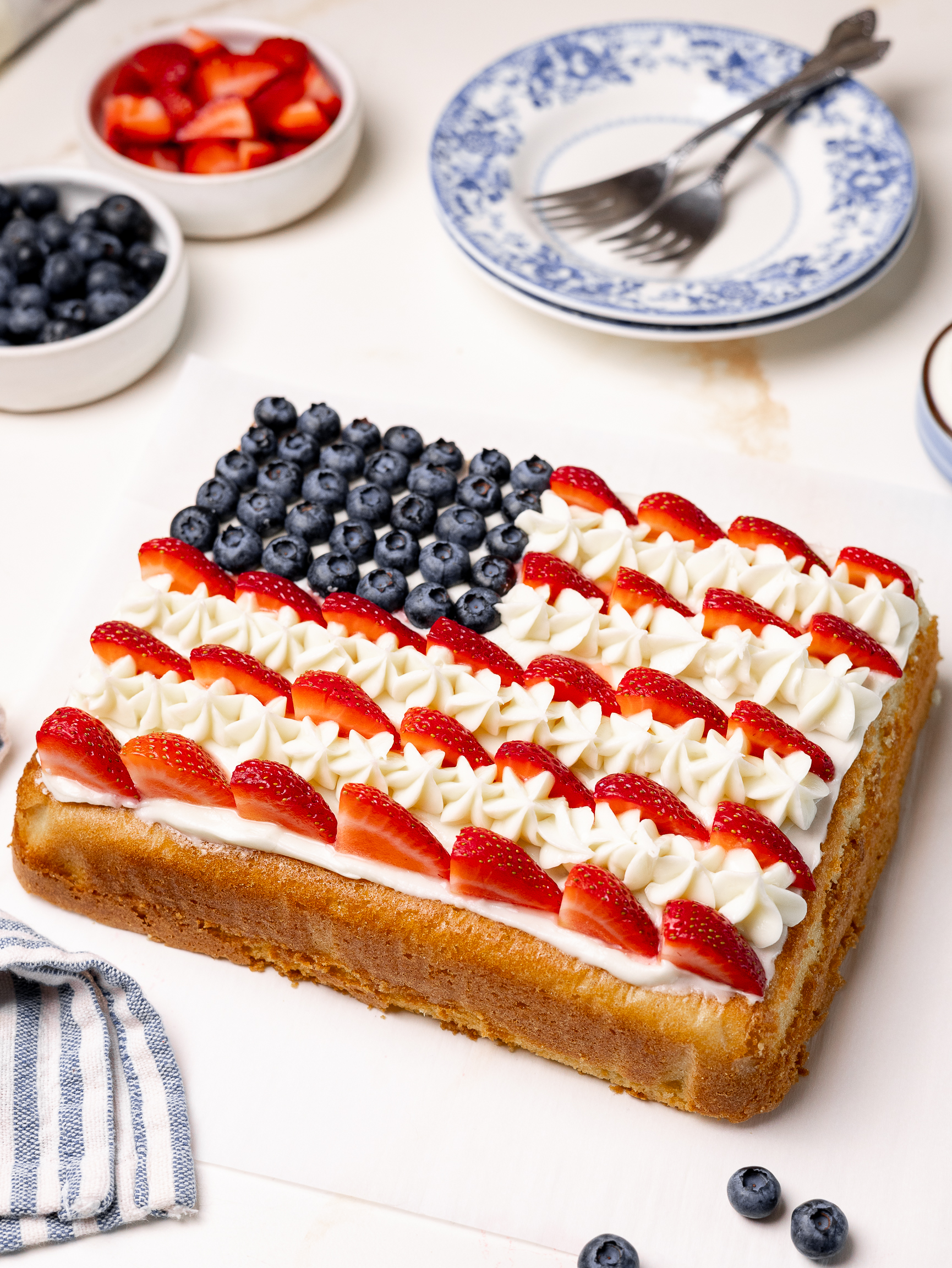 A rectangular sheet cake decorated to look like the American flag, with blueberries for the stars, piped white frosting for the stripes, and sliced strawberries for the red stripes. Surrounded by bowls of berries and empty blue floral plates with forks.