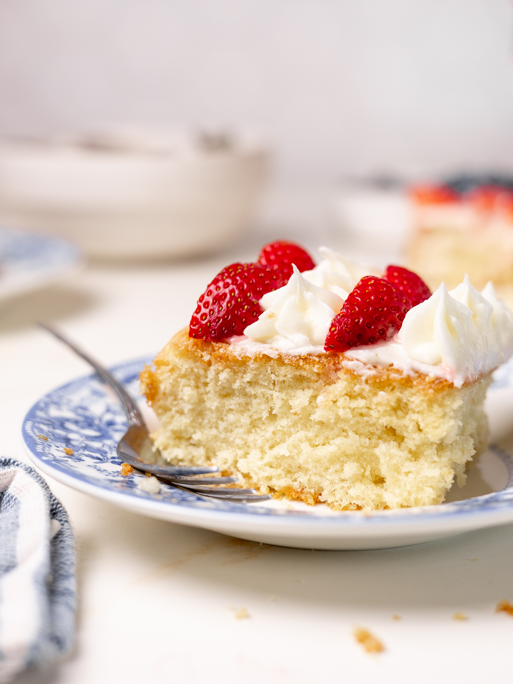 A close-up slice of vanilla American Flag cake topped with whipped frosting and fresh strawberries, served on a blue floral plate with a fork resting beside it.