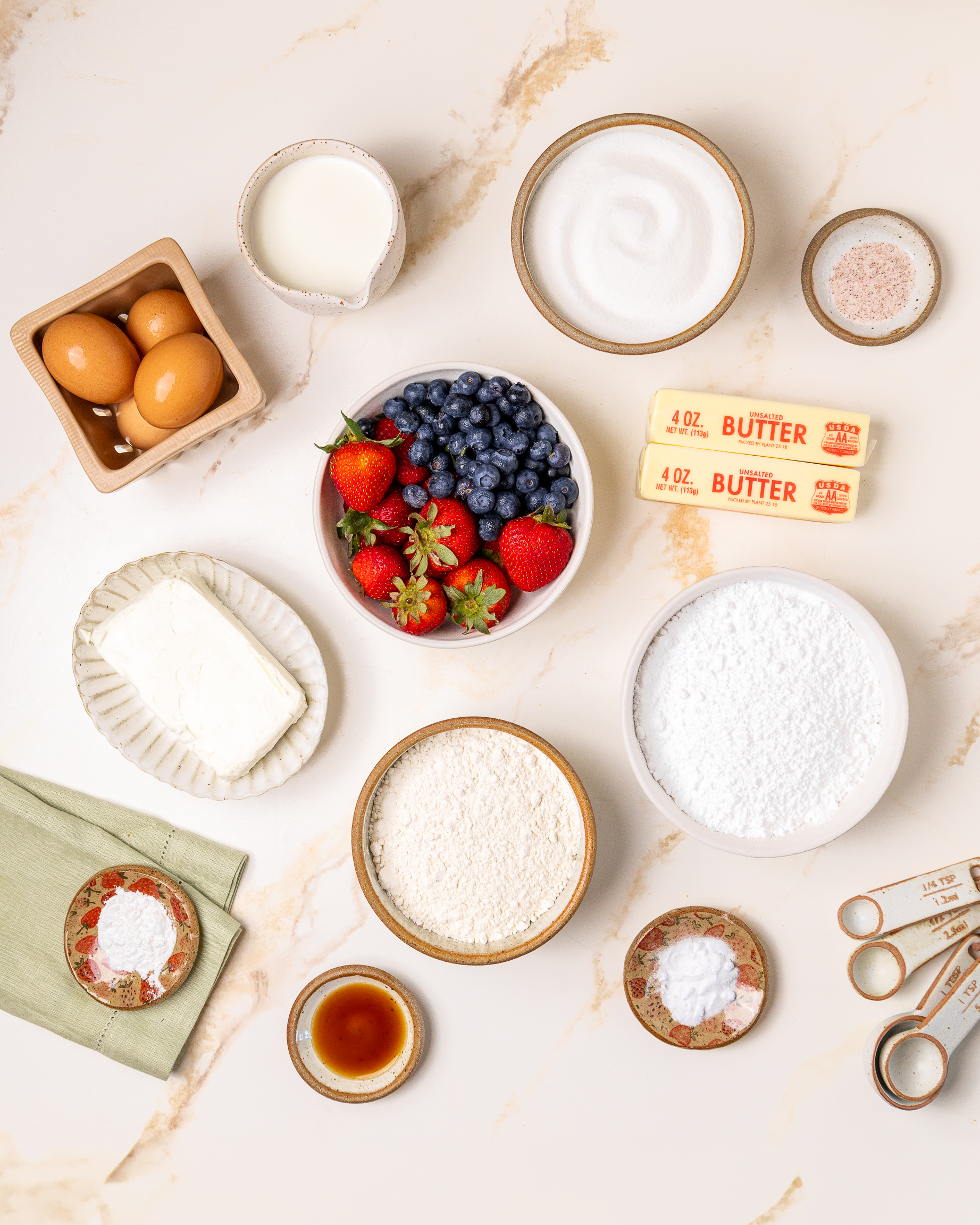 Flat lay of ingredients for an American flag cake, including strawberries, blueberries, flour, sugar, butter, eggs, cream cheese, powdered sugar, vanilla extract, baking powder, milk, and salt, arranged on a marble surface with measuring spoons.