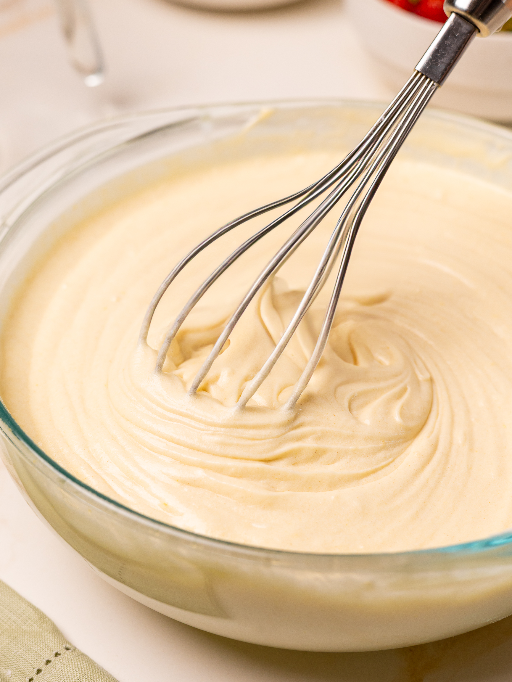 A glass bowl filled with smooth vanilla cake batter being whisked by a metal balloon whisk, showing its light and creamy texture.