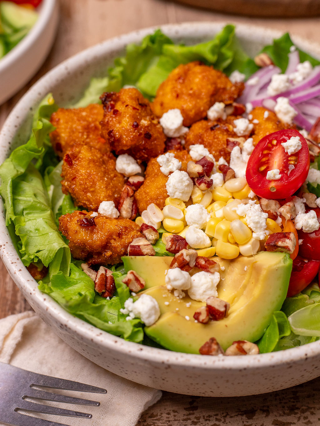 Close-up of hot honey chicken summer salad in a white bowl with feta, avocado, and toasted almonds.