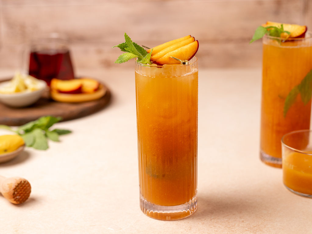 A tall glass of peach and mint iced tea surrounded by ingredients.