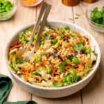 A bowl of ramen noodle salad on a rustic wooden table surrounded by ingredients.