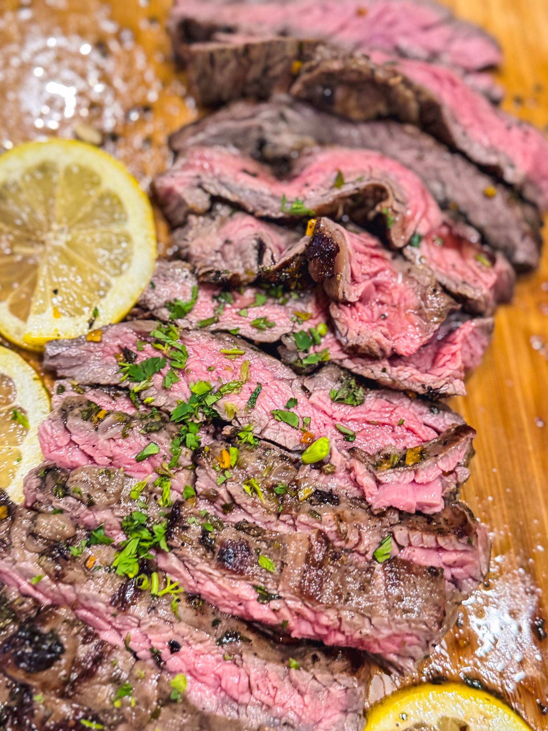 Close up of sliced skirt steak with chimichurri sauce over a wooden cutting board with a side of lemon.