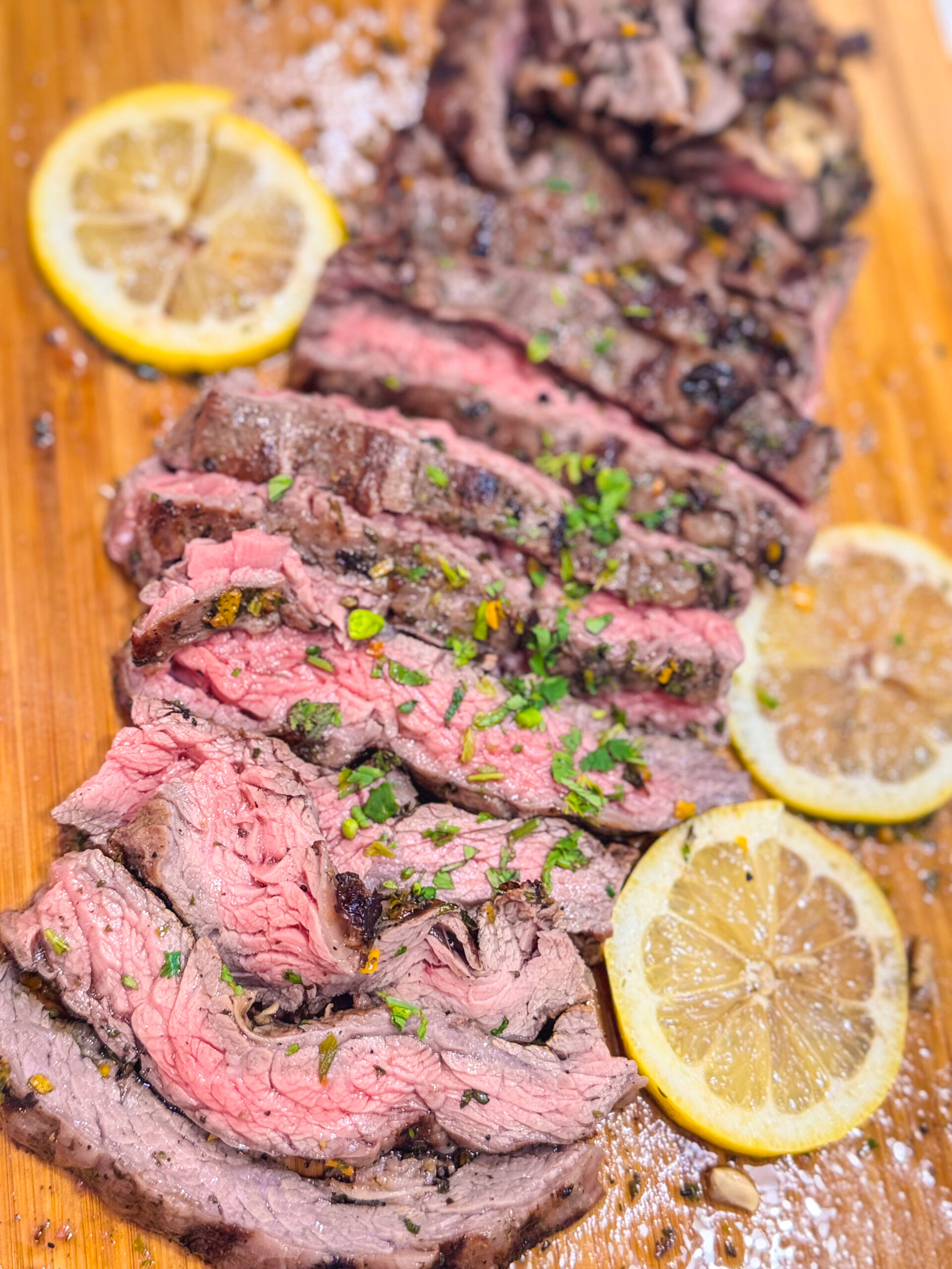 Close up of sliced skirt steak with chimichurri sauce over a wooden cutting board with a side of lemon.