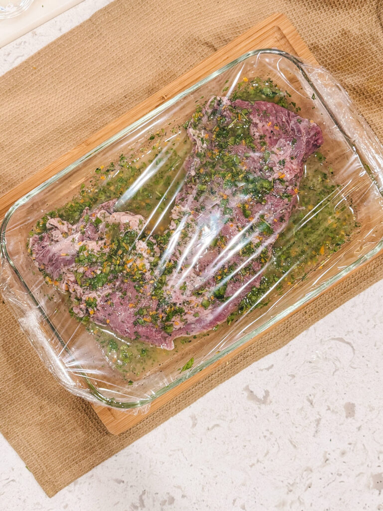 Skirt steak in a glass dish of chimichurri marinade with plastic wrap over it.
