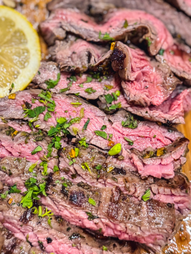 Close up of sliced skirt steak with chimichurri sauce over a wooden cutting board with a side of lemon.
