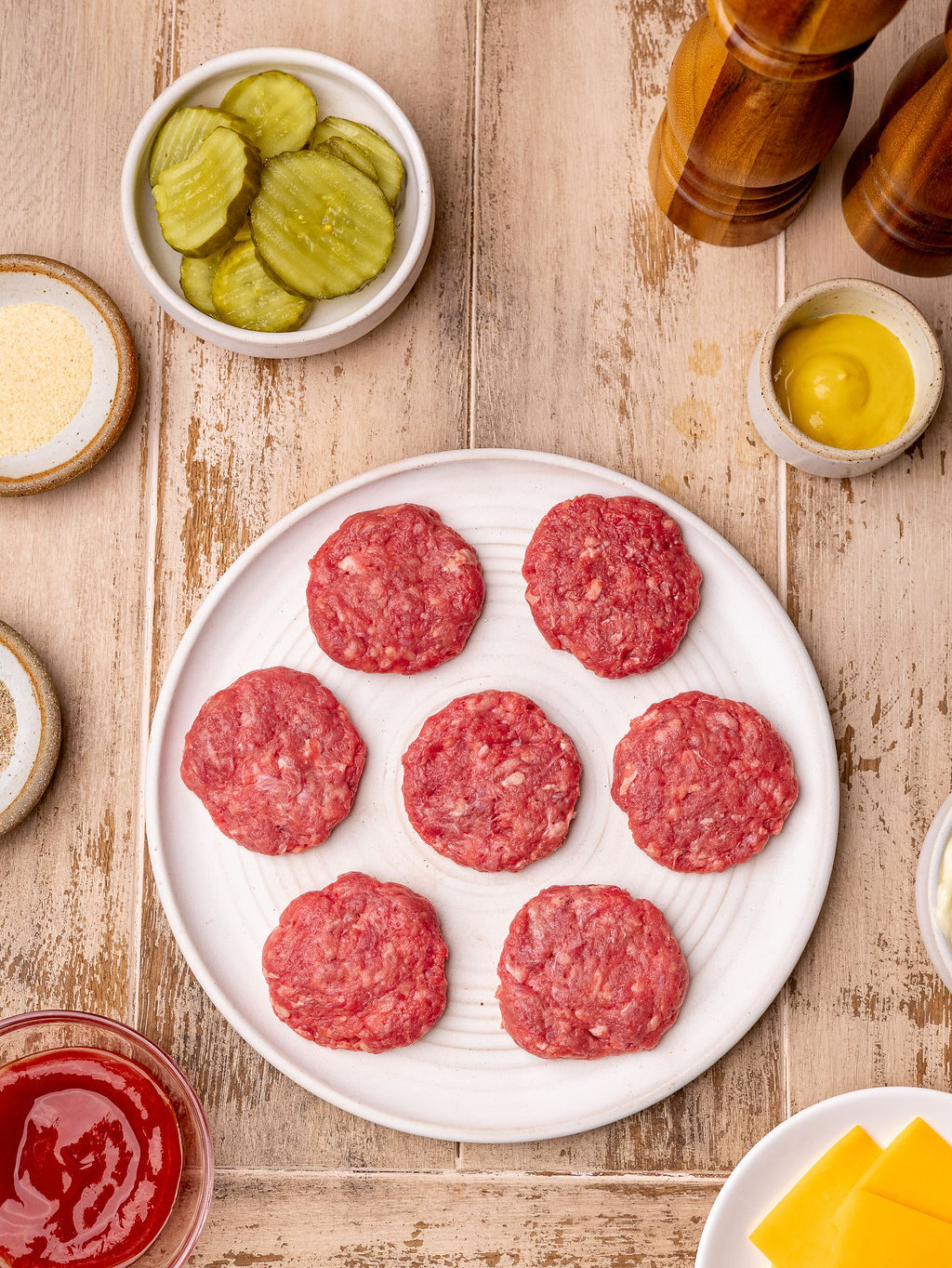 Seven burger patties on a white plate surrounded by ingredients over a rustic table.