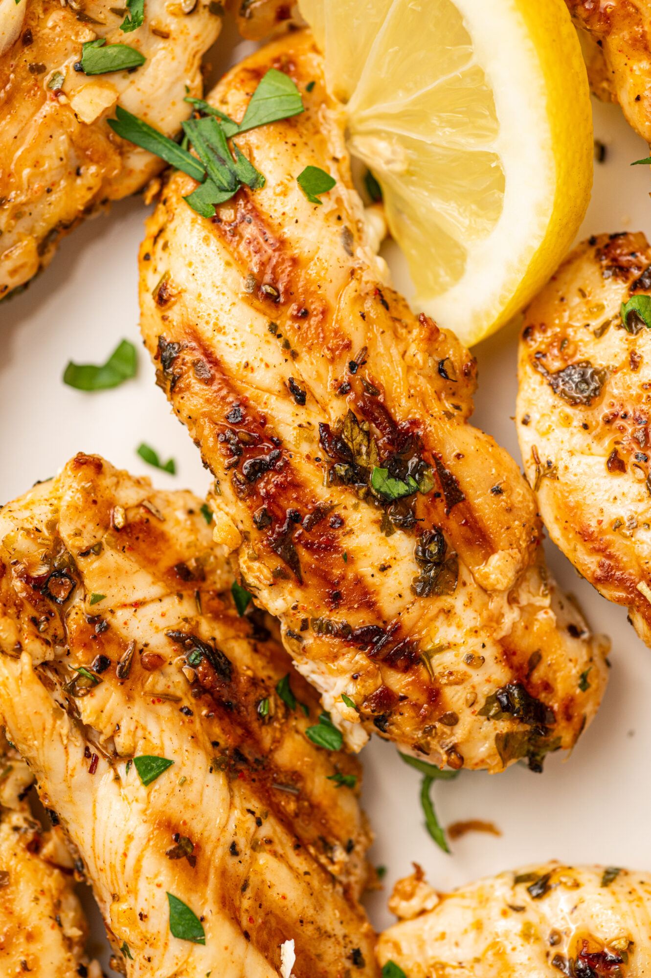 Close-up of grilled chicken tenders on a white serving plate garnished with herbs and lemon wedges.
