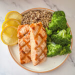 Grilled salmon with lemon butter sauce served with broccoli, lemon slices, and wild rice.