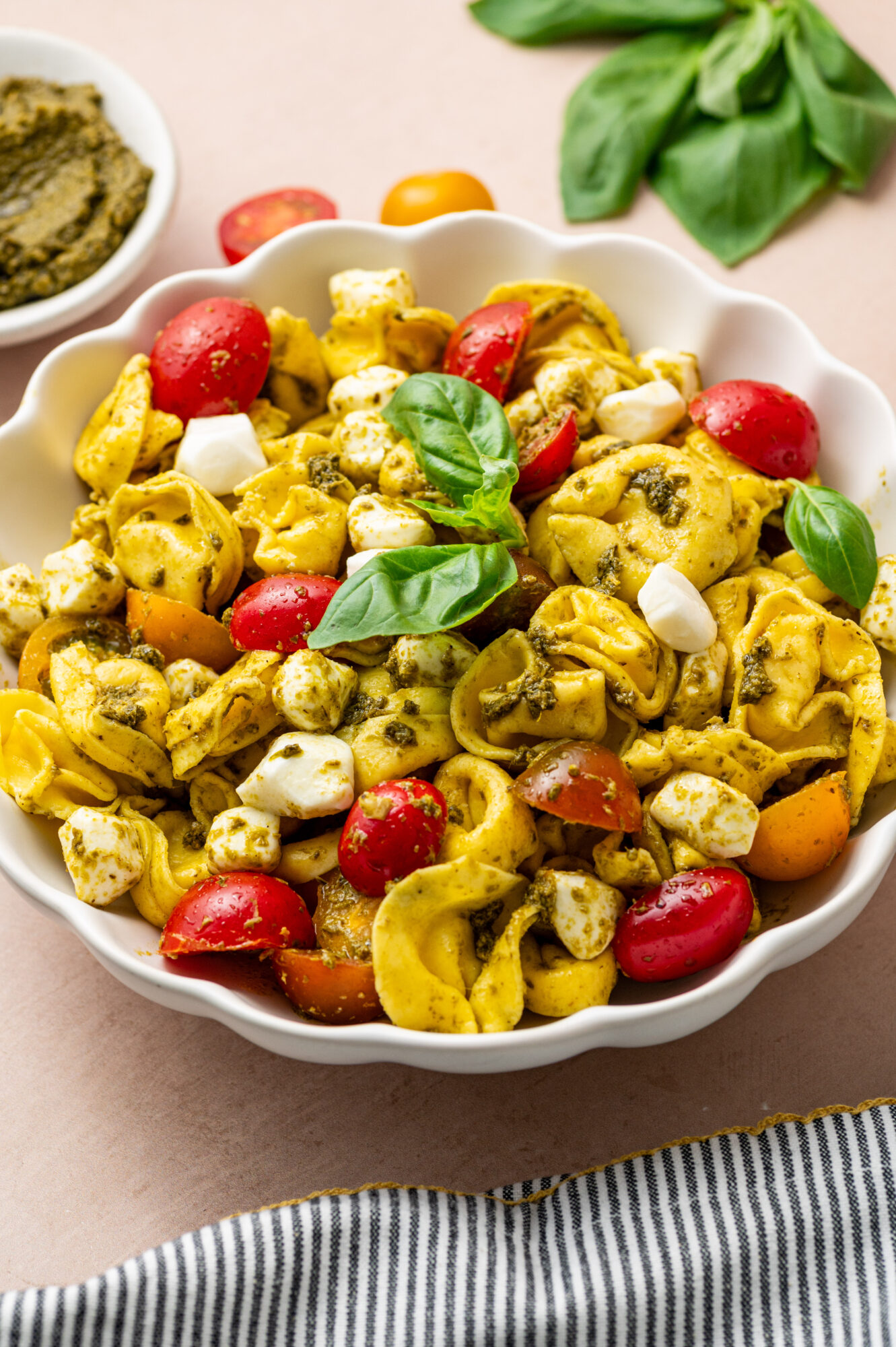 A white bowl of pesto tortellini salad with tomato, mozzarella, and basil.